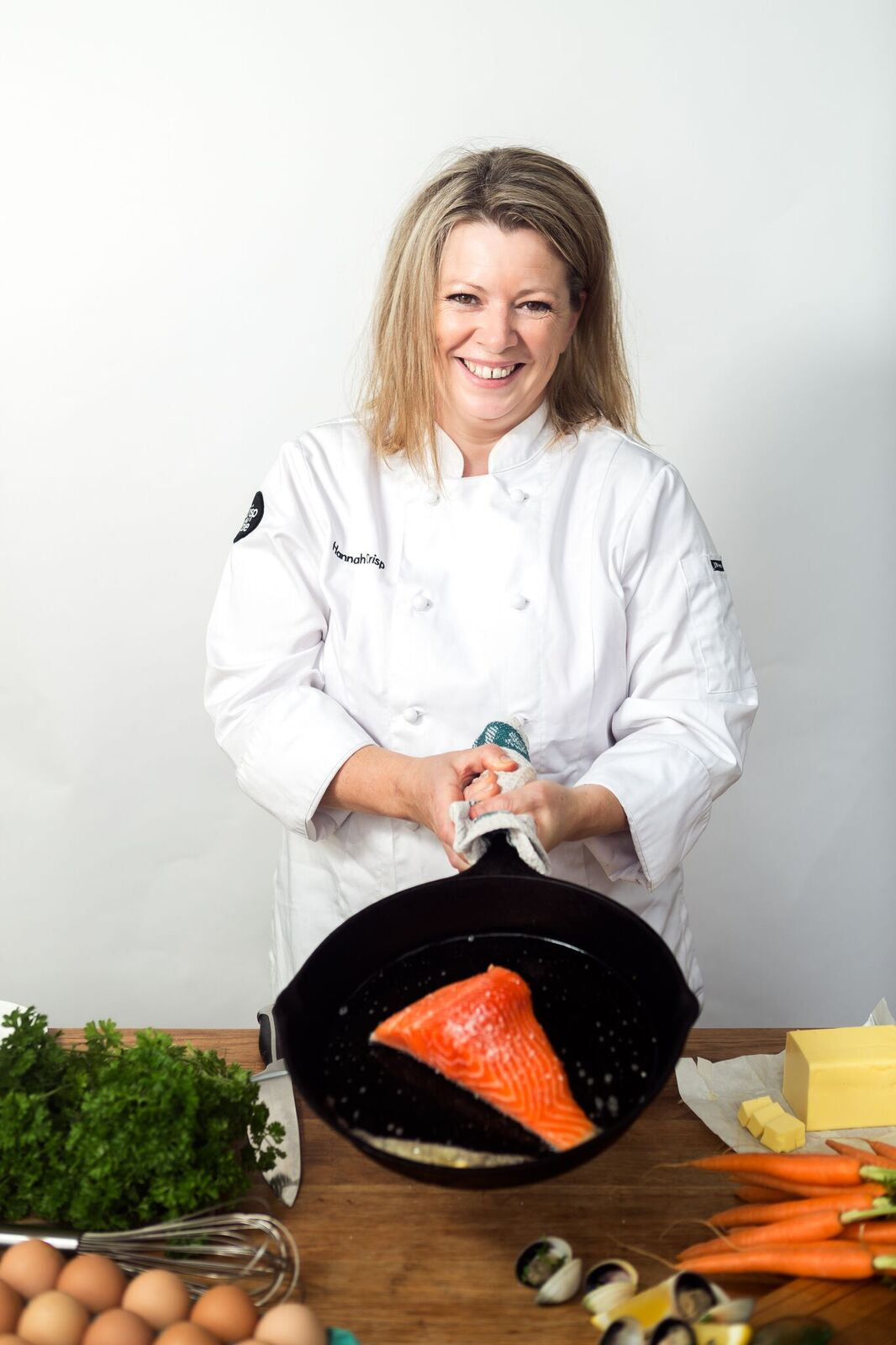 Smiling chef in white uniform holding a skillet with a salmon fillet, surrounded by fresh ingredients on a wooden table