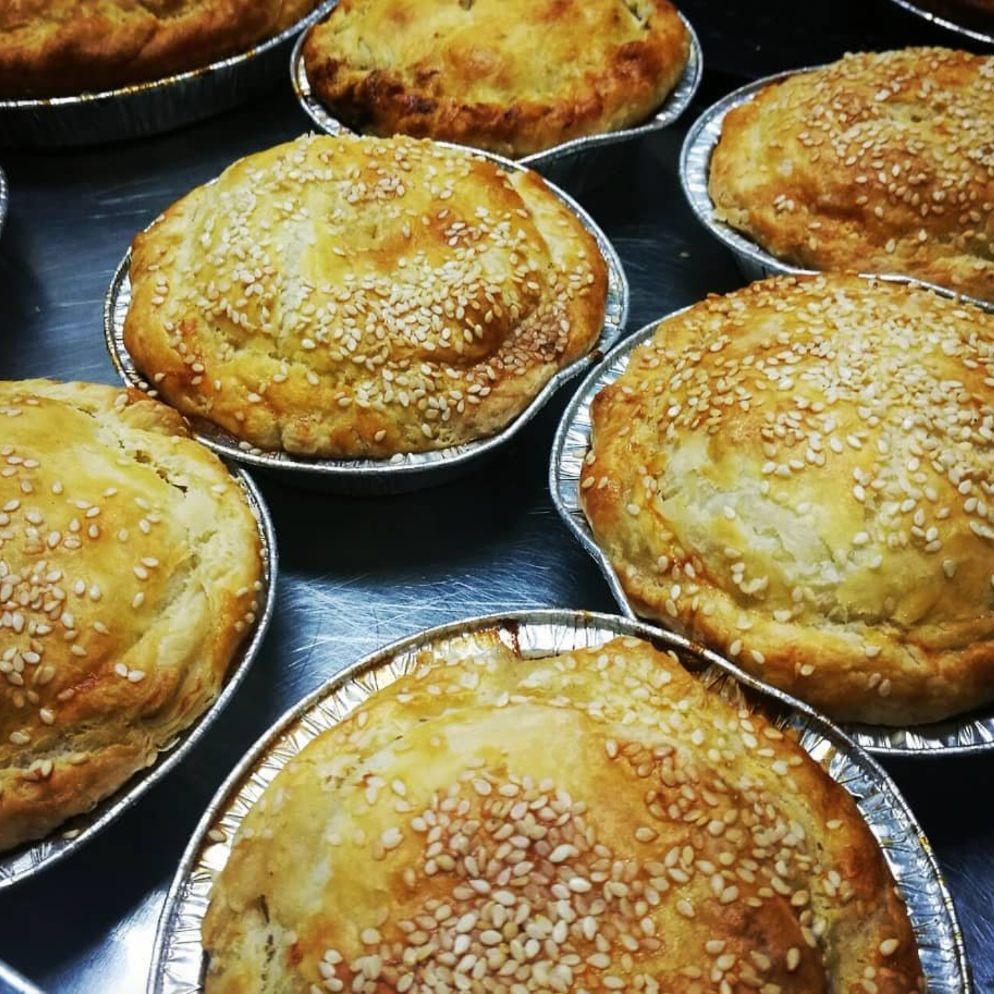 Picture of several beef and Ale pies that have just been cooked. Golden pastry and sesame seeds on top. sitting on bench in tin foil casings