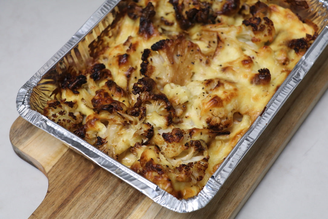 Cooked cauliflower cheese ready meal in tin foil container sitting on a wooden serving board. White background