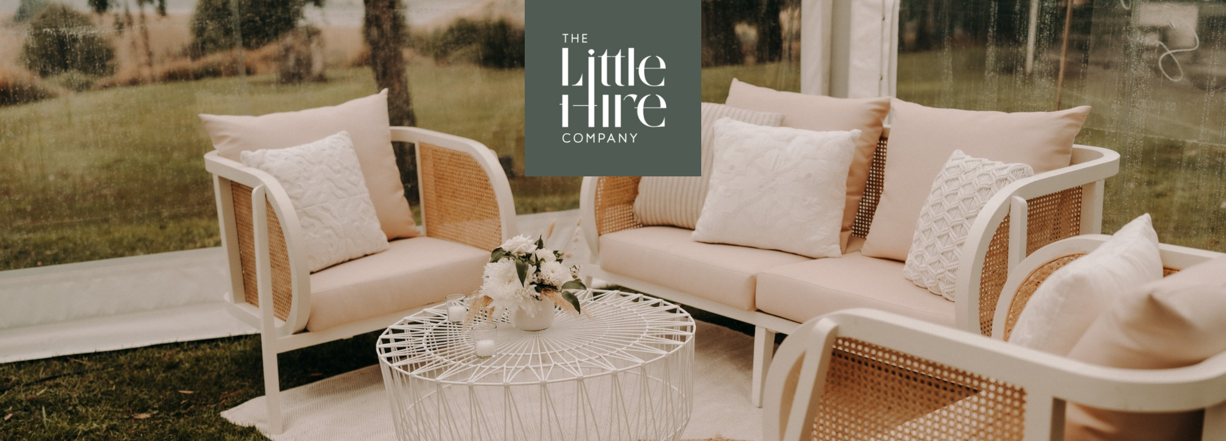 Elegant outdoor lounge setup with cream sofas, textured pillows, white wire coffee table, and fresh floral centrepiece under a clear marquee