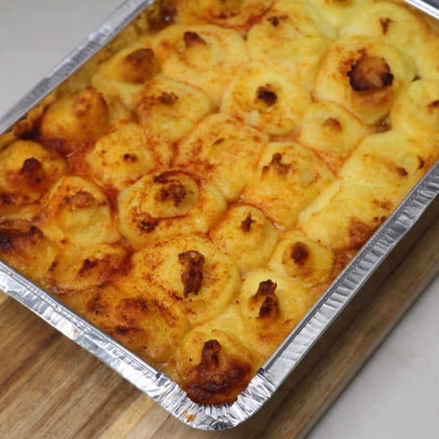 Cooked Shepards Pie ready meal in tin foil container sitting on a wooden serving board. White background