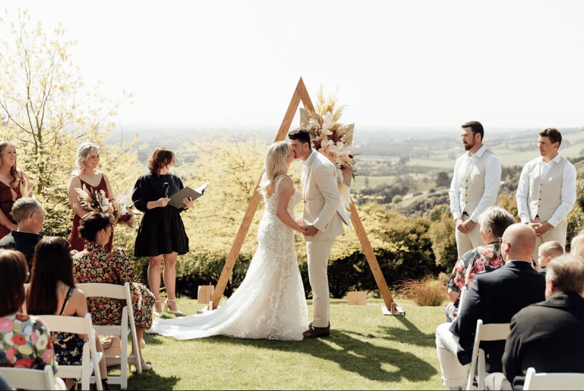 Bride and groom kissing at outdoor wedding ceremony with wooden arch and floral arrangement, surrounded by guests.